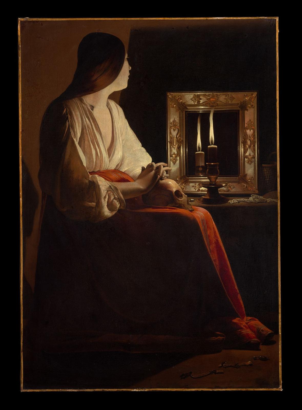 A young woman in a white blouse and red skirt sits in a dark room, one hand resting on a skull in her lap, looking at a candle flame and its reflection in a gilt-framed mirror.