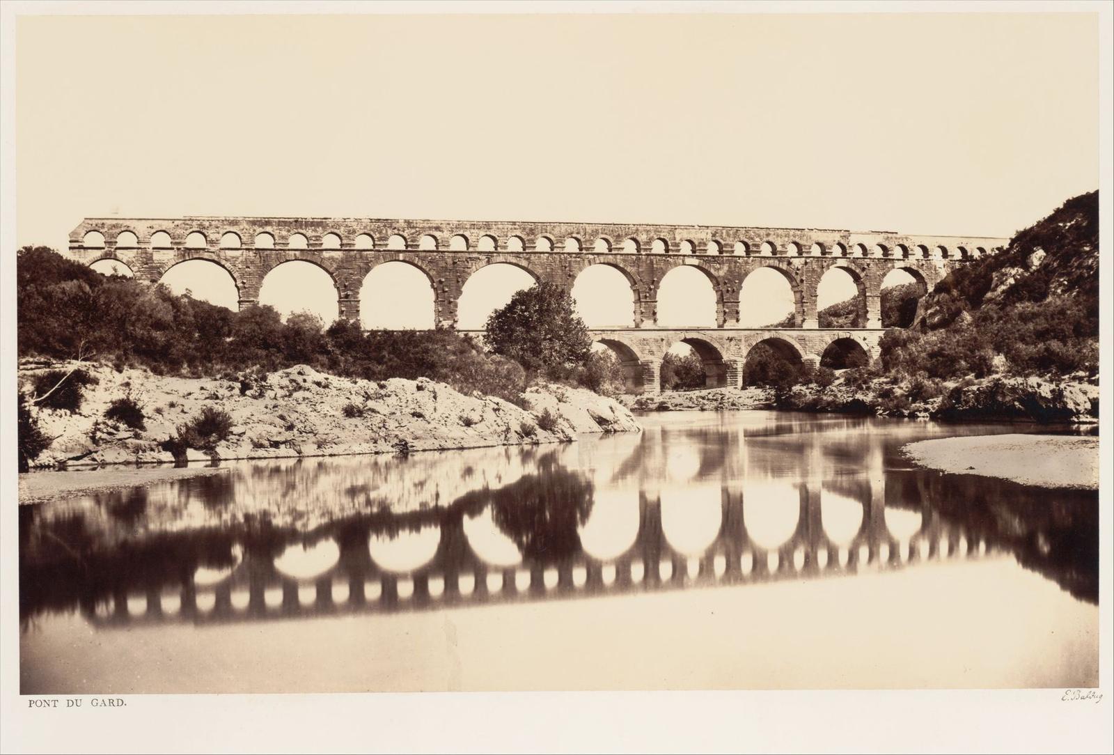 An early photograph of the Pont du Gard, a Roman aqueduct bridge with three tiers of stone arches spanning a calm river, its arches reflected in the water below.