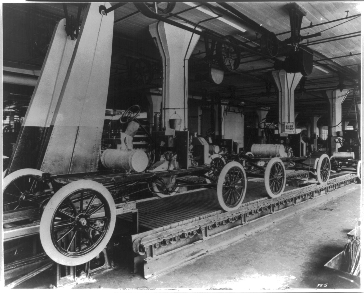 A black-and-white photograph of a factory floor with a long raised conveyor carrying a row of car chassis with spoked wheels and fuel tanks, under overhead belts, pipes and hanging parts.