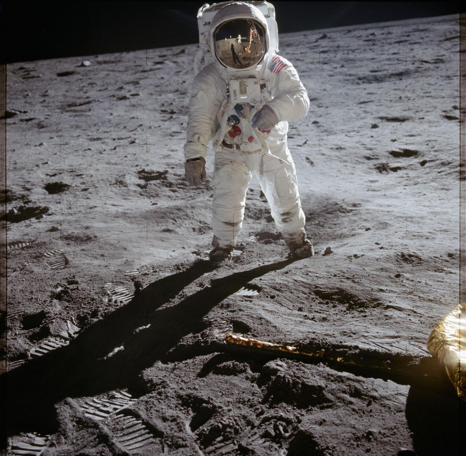 An astronaut in a white spacesuit stands on grey lunar soil marked with bootprints; his gold visor reflects the photographer and the lunar module.