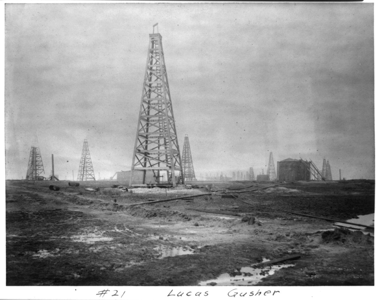 A black-and-white photograph of a tall wooden oil derrick on muddy, puddled ground, with more derricks and a large round storage tank behind it under a grey sky; a handwritten label at the bottom reads "#21 Lucas Gusher".