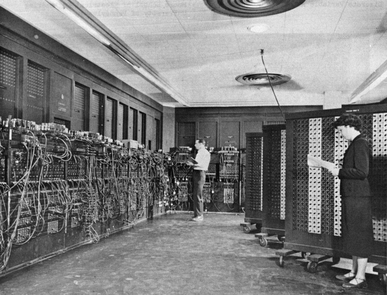 A long room lined with tall cabinets covered in switches and cables; a man stands at the far panels and a woman in the foreground sets switches on a wheeled unit.