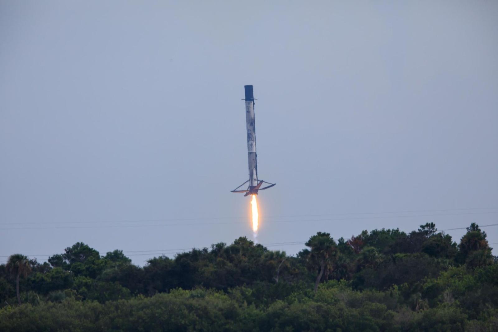 A tall white and black rocket stage descends tail first on a bright orange flame over a line of green trees, its four legs spread beneath it.