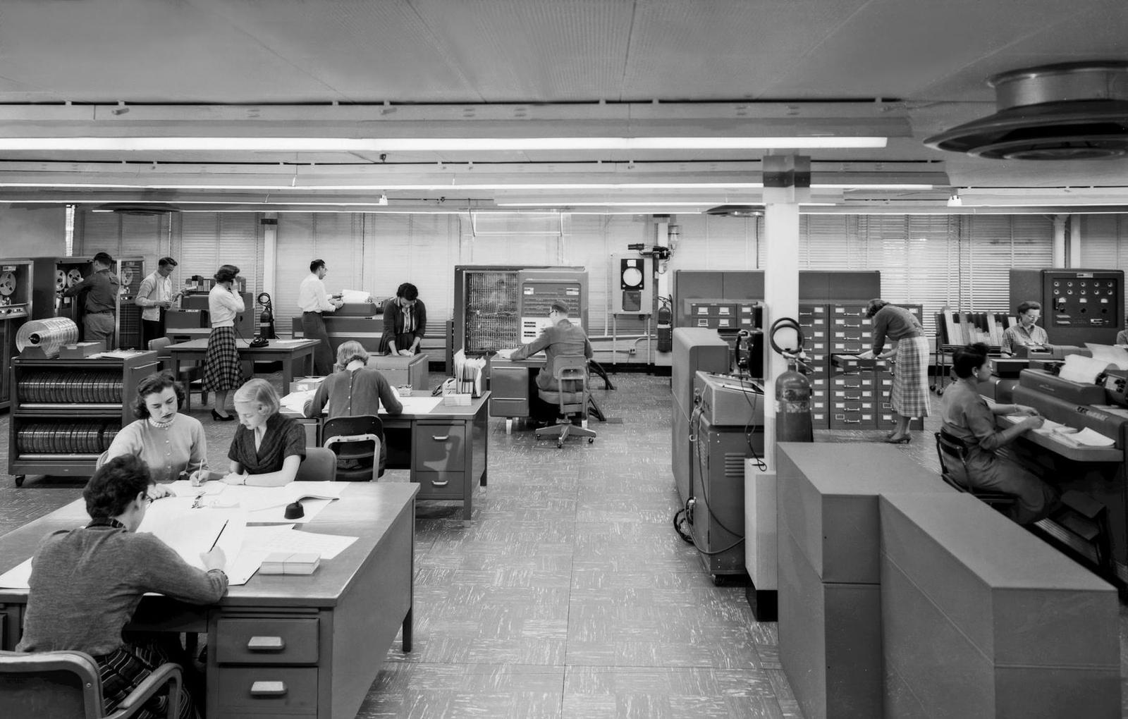 A black-and-white photograph of a large open office in the 1950s, where women and men sit at desks with papers and stand at the tall grey cabinets and tape drives of an early computer.
